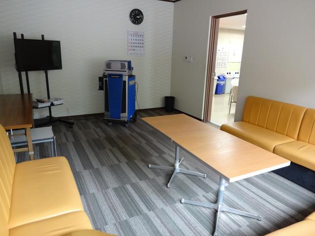 グレーの床に新しい机にソファーとテレビ、青い台の機械が置かれた綺麗な部屋の写真