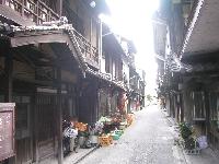 路地の両脇に昔ながらの2階建ての木造の家が並んでおり、左側の店先に花や商品が並んでいる木江の町並みの写真
